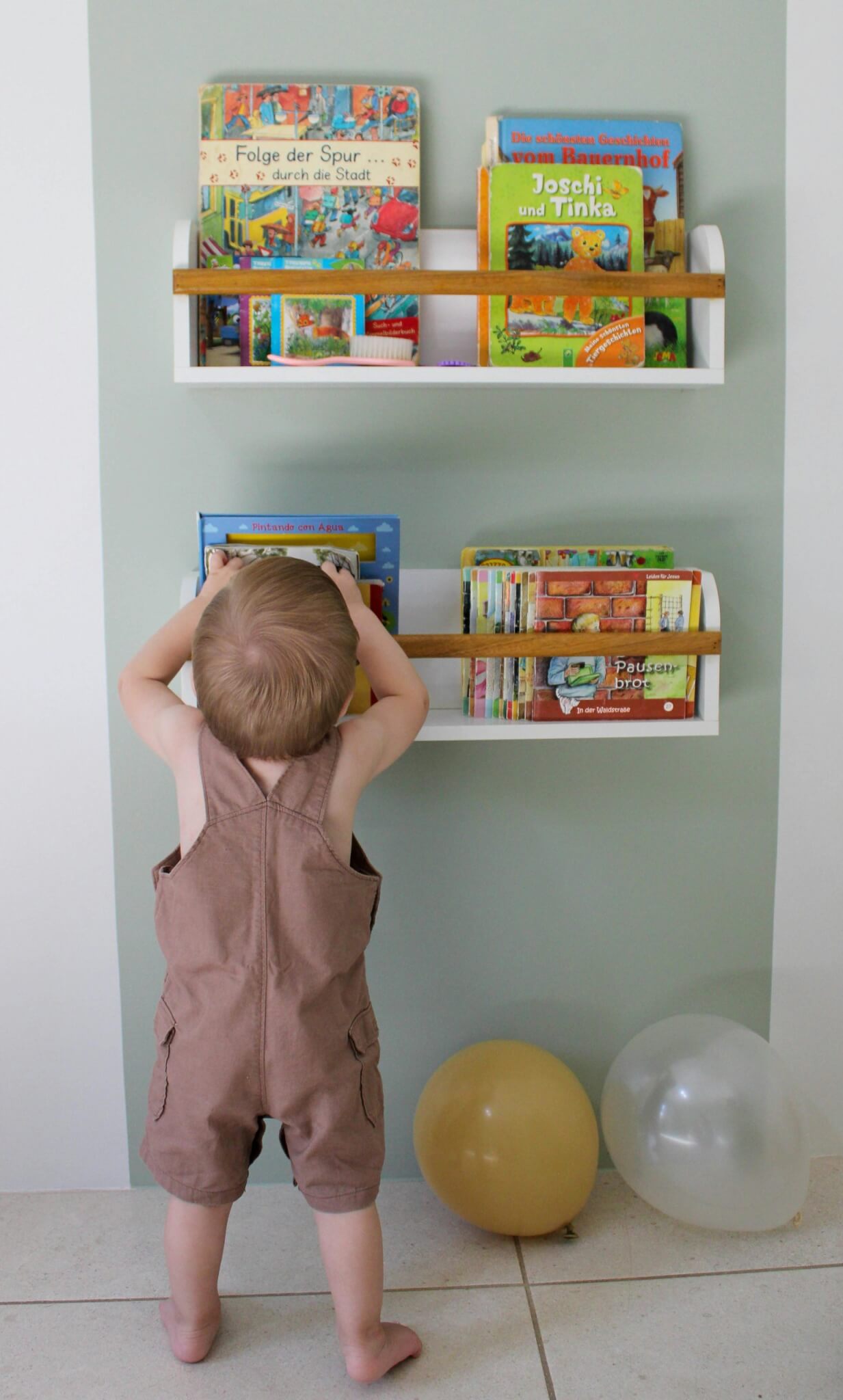 A toddler stands reaching for books on a wall shelf, surrounded by colorful balloons.