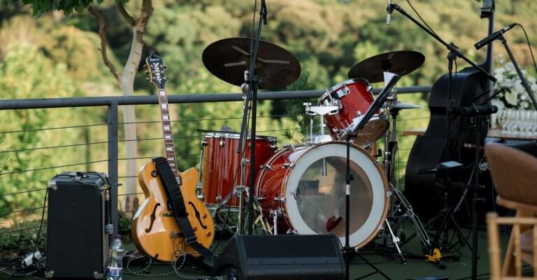 An outdoor stage setup with a drum set, guitar, and amplifier amidst greenery.
