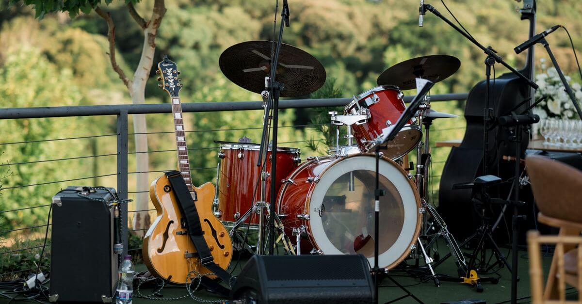 An outdoor stage setup with a drum set, guitar, and amplifier amidst greenery.